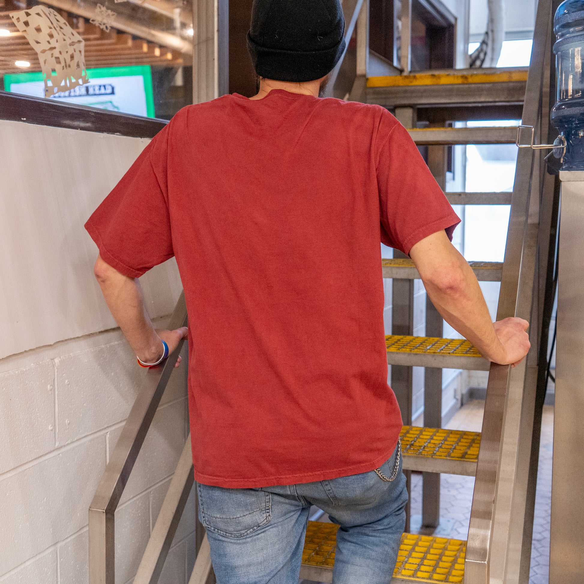 Someone wearing an Inn Red Line Art Tee from GO WEST T-SHIRT COMPANY, jeans, and a black beanie is seen from behind walking up an indoor metal staircase with yellow safety strips.