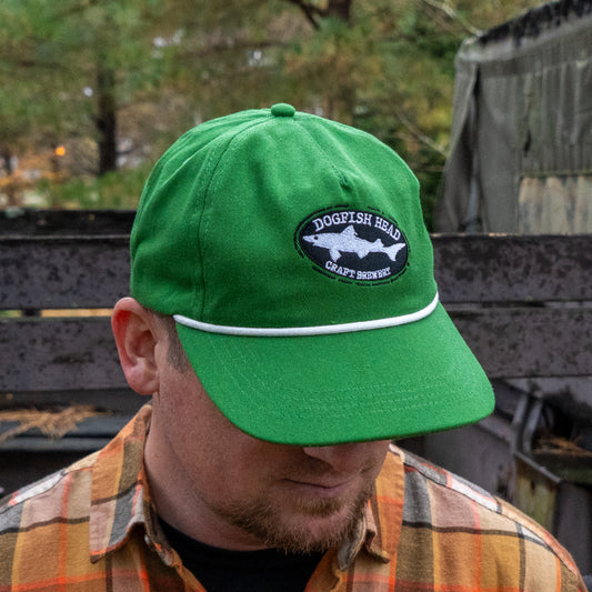 A person wearing a Merchtank Kelly Green Rope Hat with a snapback closure and a plaid shirt looks down outdoors, with trees and metal structures in the background.
