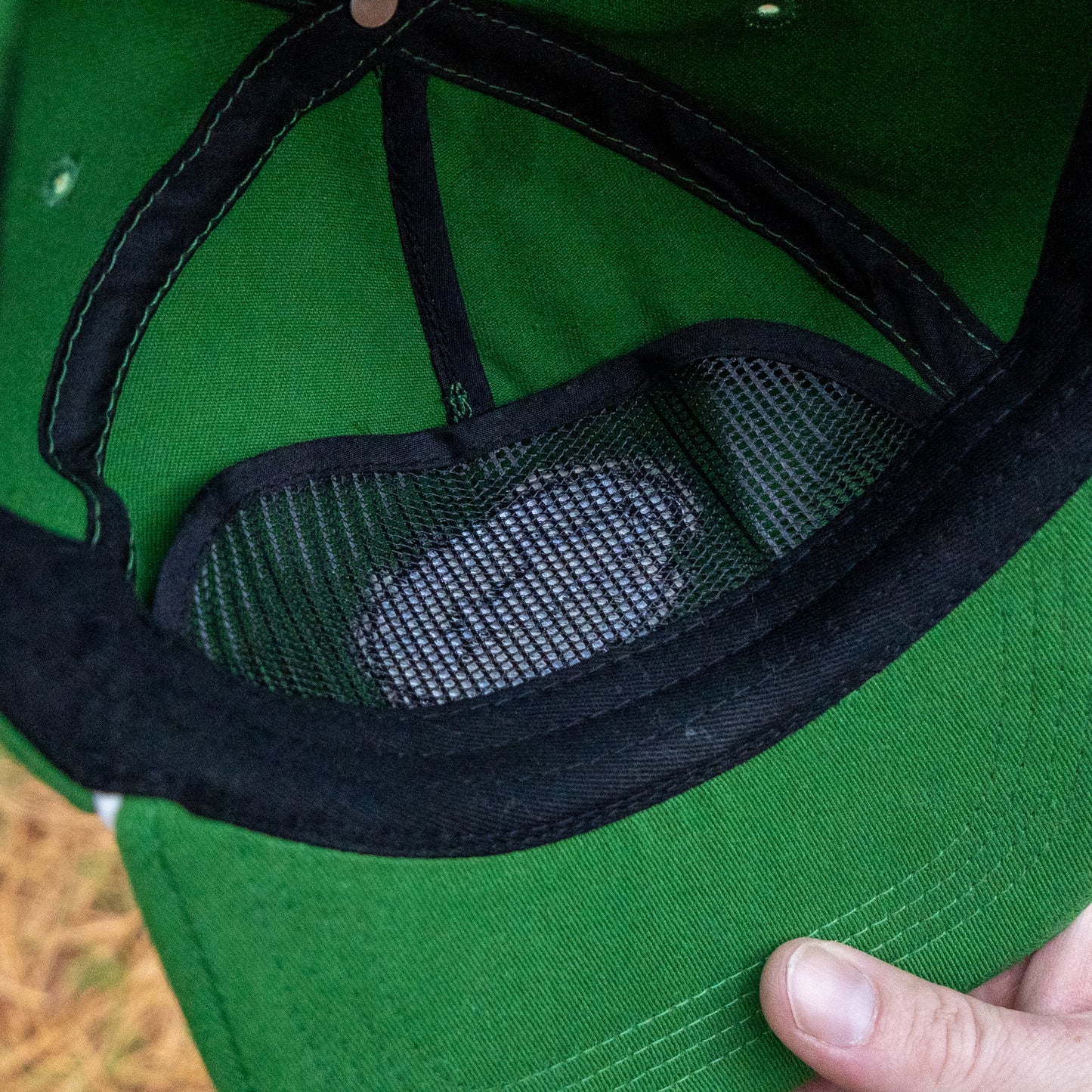 Close-up of the inside of a Merchtank Kelly Green Rope Hat, showing a white object in a black mesh pocket. A hand holds the brim of this one-size-fits-most cap.
