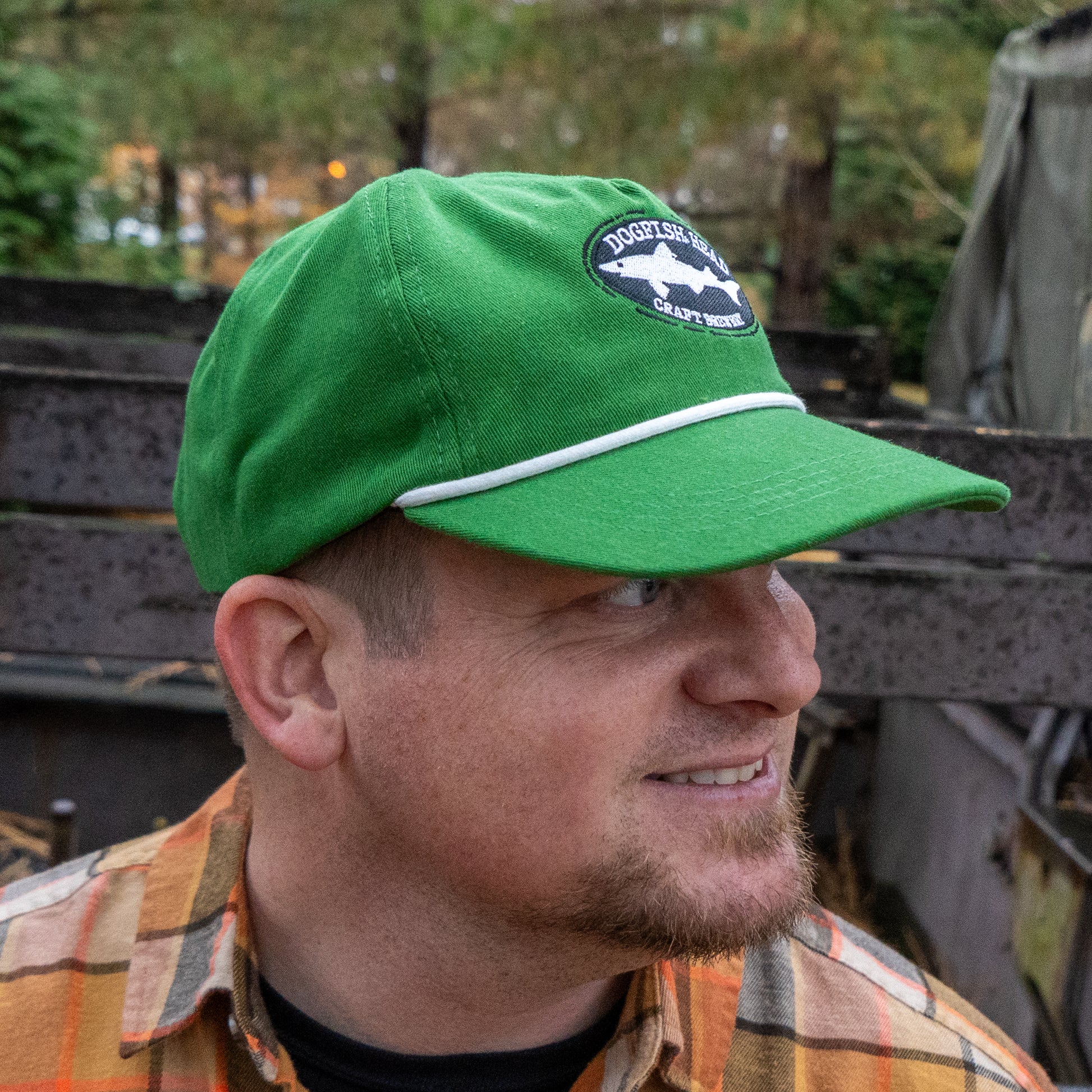 A man in a Merchtank Kelly Green Rope Hat with a patch and snapback smiles, looking right. He wears a plaid shirt and has a short beard, standing outdoors among trees and rustic metal structures.