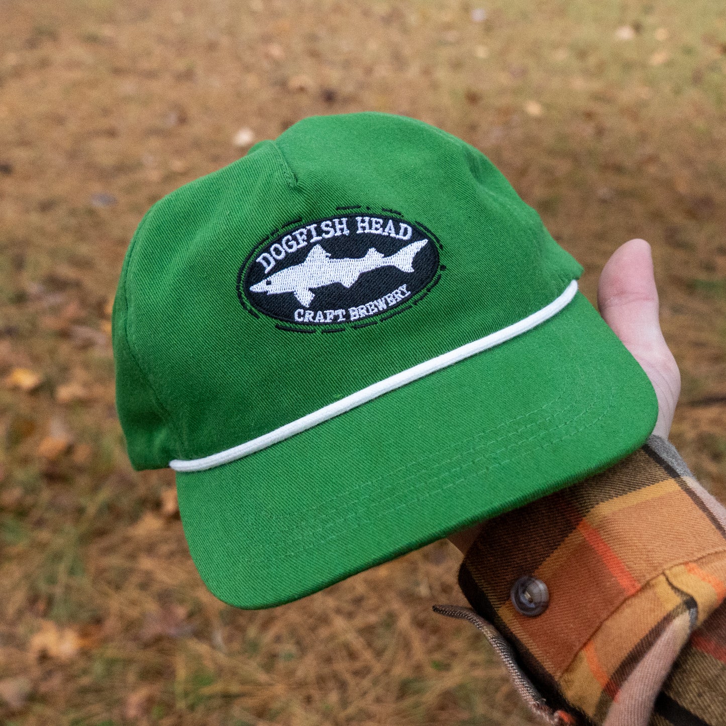 A hand holds a Merchtank Kelly Green Rope Hat with a snapback closure outdoors on brown grass. The person is wearing a plaid shirt.