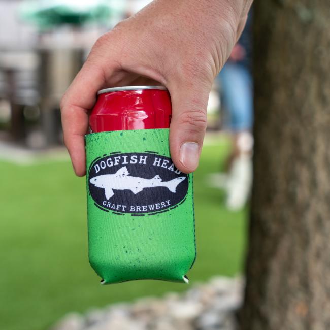 A hand holds a red can in a Merchtank Lime Green Craft Brewery 12oz Can Cooler, featuring an outdoor background with grass, rocks, a tree trunk, and blurred seating.