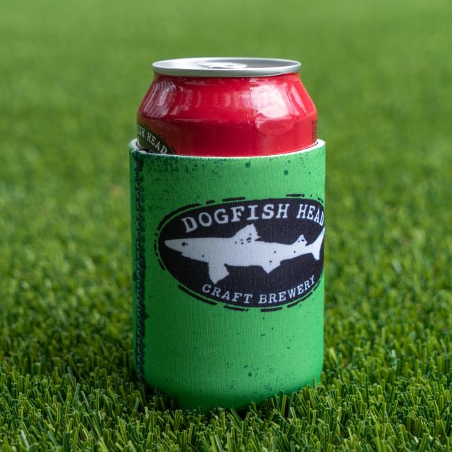 A red can sits on green artificial grass in a Lime Green Craft Brewery 12oz Can Cooler from Merchtank, featuring the Dogfish Head logo.