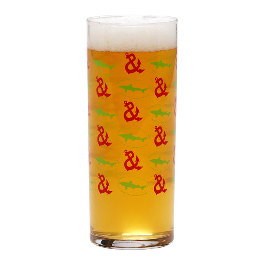 A tall glass of light amber beer with a foamy head, served in a Grandstand Glassware & Apparel Lime and Pink Collins Glass decorated with a pattern of red anchors and green sharks.