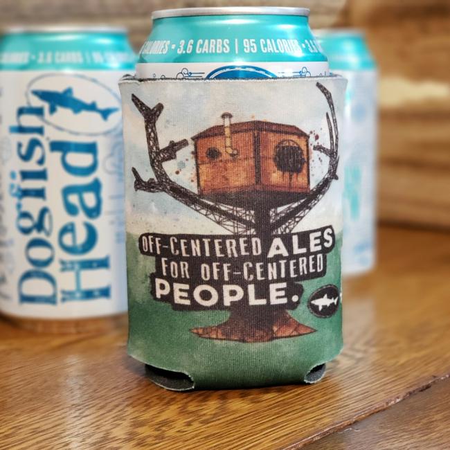 A Dogfish Head 12oz can sits in a Grandstand Glassware & Apparel Off Centered Ales Treehouse Can Cooler, which displays a treehouse and the phrase "Off-centered ales for off-centered people." Two more cans are blurred in the background.