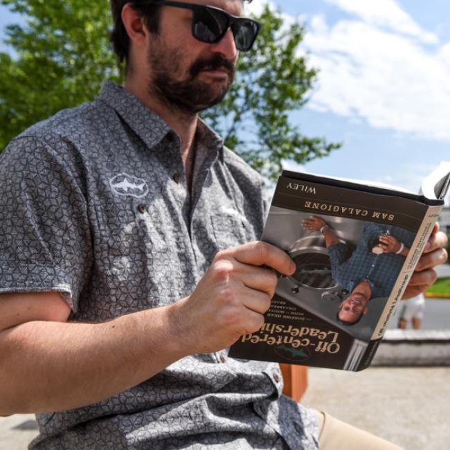 A man in sunglasses and a patterned short-sleeve shirt sits outside on a sunny day, reading "Off-Centered Leadership" by J Wiley and Sons Publishing, finding business inspiration among trees beneath a blue sky.