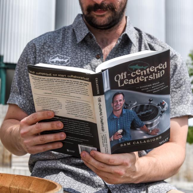 A mustached man sits outdoors reading Off-Centered Leadership by J Wiley and Sons Publishing, as shown by the cover featuring a smiling man with a glass near brewing equipment.