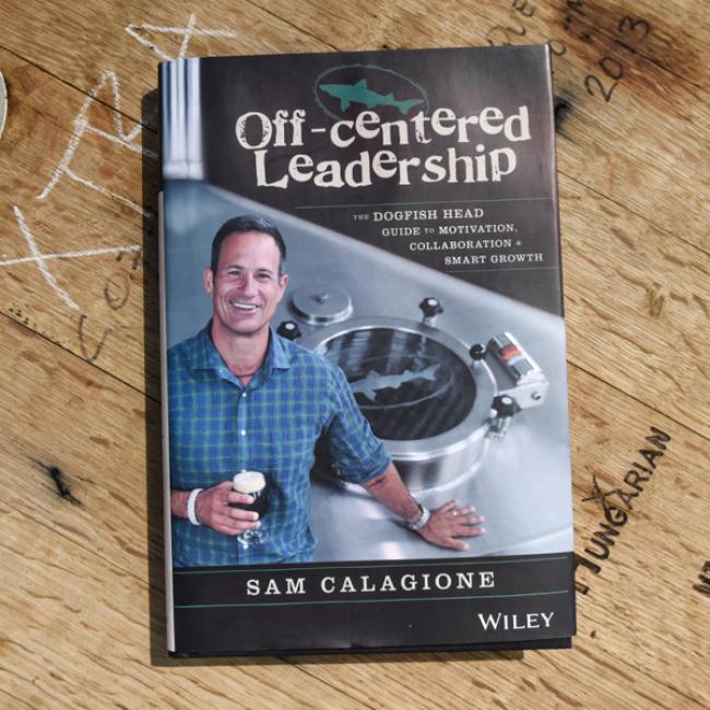 Off-Centered Leadership by J Wiley and Sons Publishing is displayed on a wooden table. The cover shows the author smiling with a beer in hand, brewing equipment behind him, emphasizing themes of leadership and collaboration.