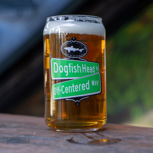 An amber beer sits on a wooden surface in the Off Centered Way Can Glass by Grandstand Glassware & Apparel, featuring Dogfish Head branding and green street sign-style labels celebrating 30 years. The background is blurred.