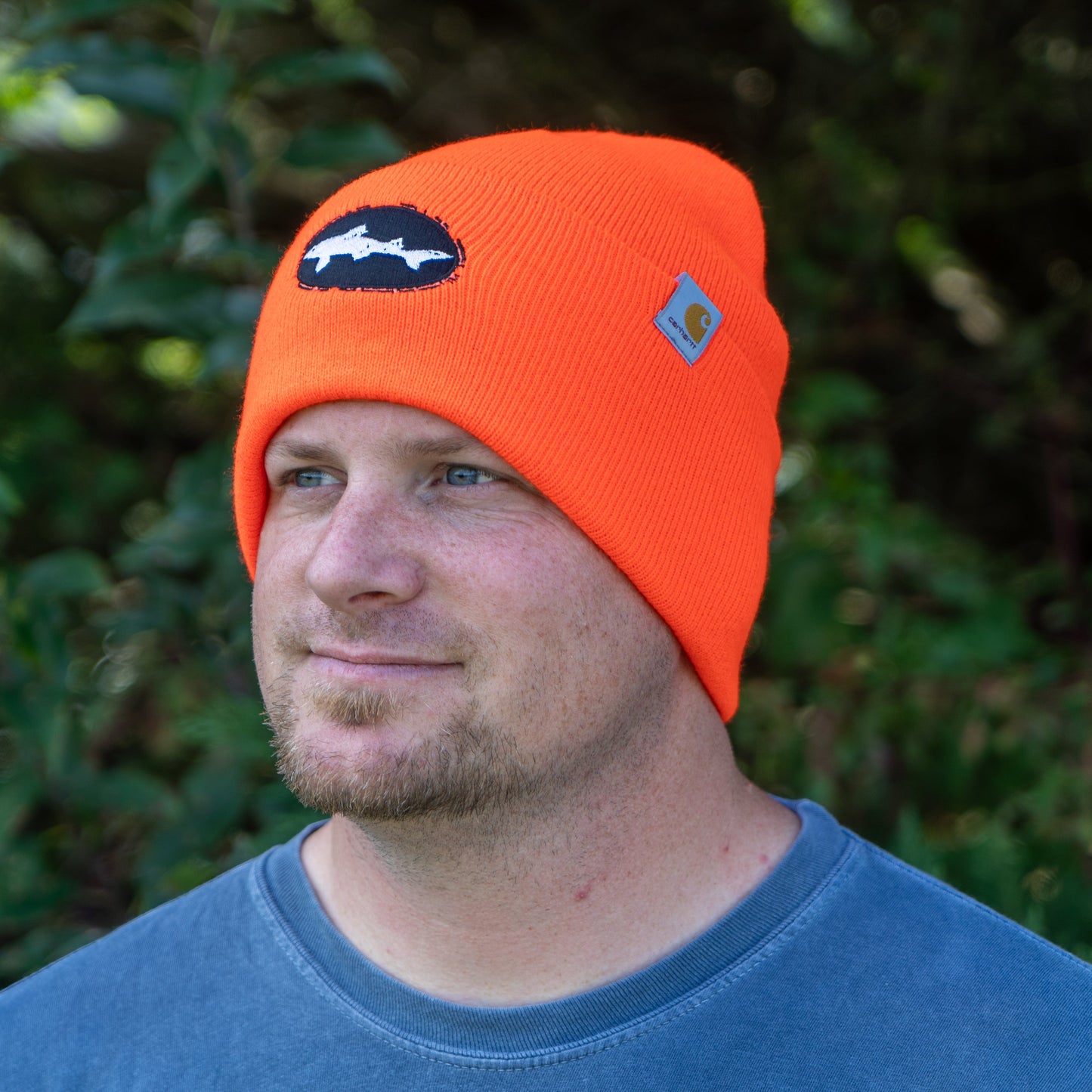 A man stands outdoors amid green foliage wearing the Orange Carhartt Beanie by Mirror Image Inc., which features a Dogfish Head patch and logo, pairing it with a blue shirt for warmth and style.
