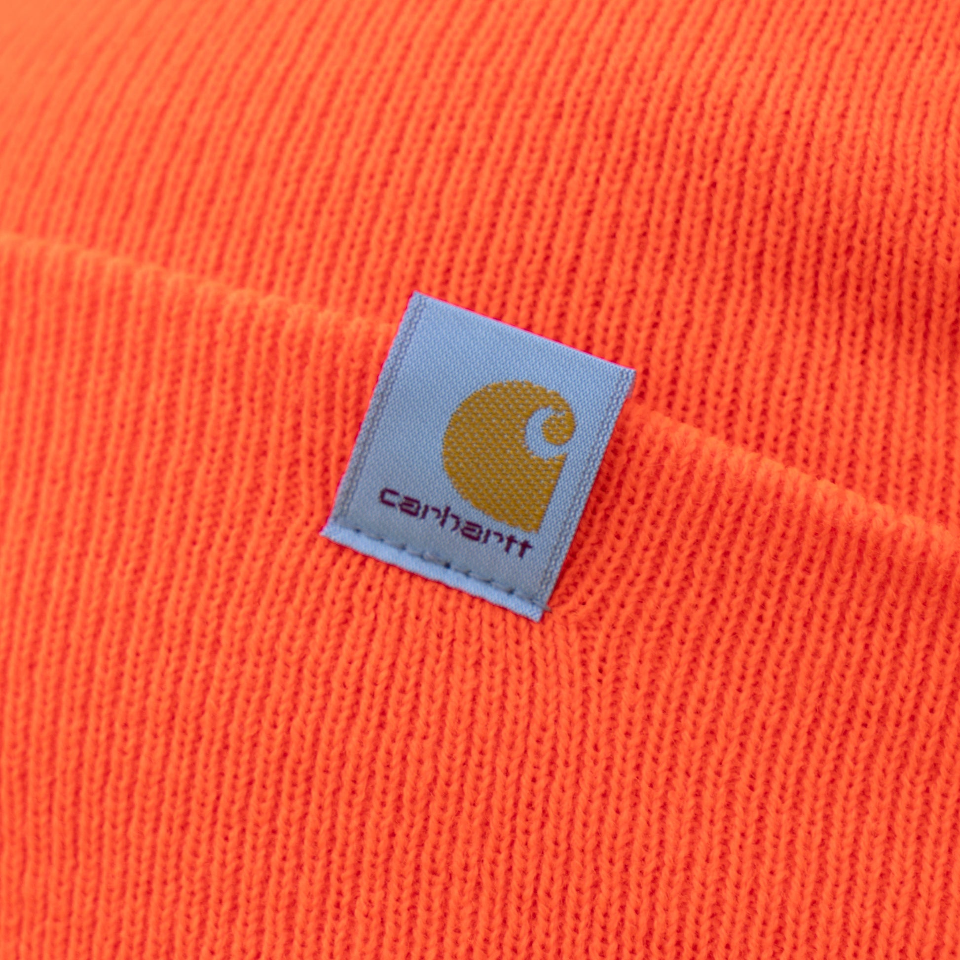 Close-up of an Orange Carhartt Beanie by Mirror Image Inc., showcasing its knit fabric and sewn-on patch with a yellow C and gray brand name, designed to keep your head warm.
