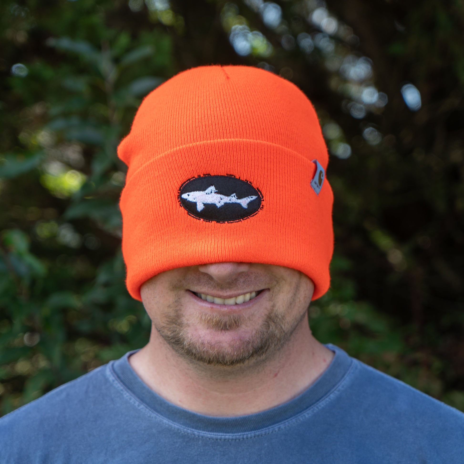 A man smiles outdoors in a bright Orange Carhartt Beanie by Mirror Image Inc., pulled over his eyes and featuring a shark patch against green foliage—perfect for keeping your head warm on cool days.