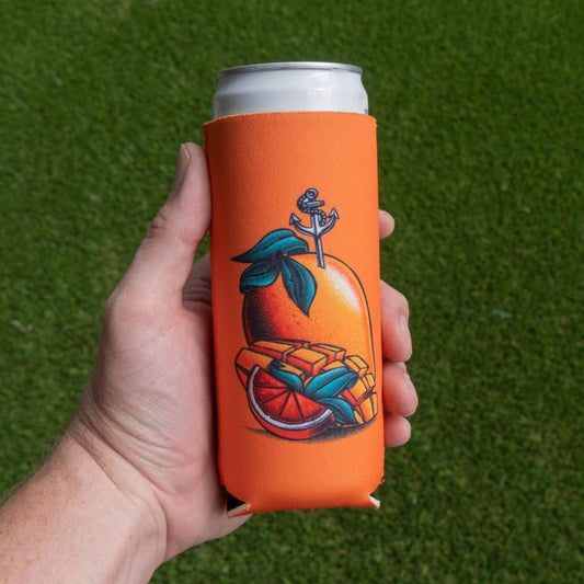 A hand holds a Merchtank Orange Mango Slim Can Cooler, decorated with orange slices, mango, and an anchor around a tall canned cocktail. Lush green grass is seen in the background.
