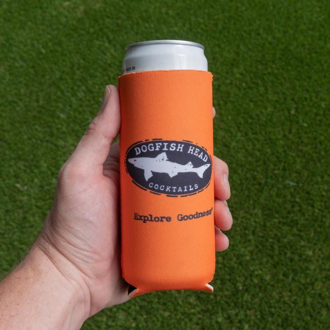 A hand holds a white canned cocktail wrapped in a Merchtank Orange Mango Slim Can Cooler, featuring the logo and "Explore Goodness" text, set on green grass.