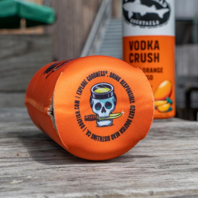 A Merchtank Orange Mango Slim Can Cooler with a skull and cocktail design rests on wood, while blurred cans of Dogfish Head Vodka Crush appear in the background.