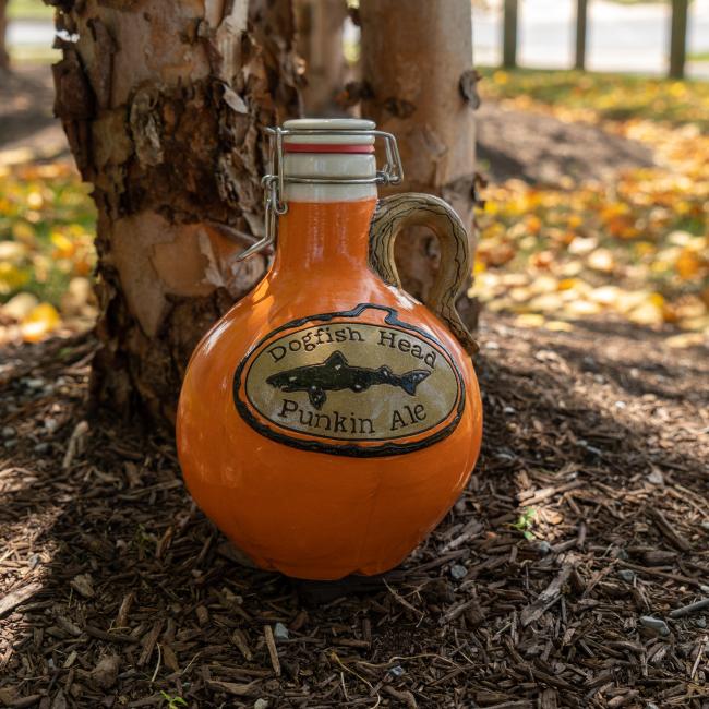 An Orange Punkin Growler by Romanick Pottery rests on mulch at a tree's base, surrounded by fallen autumn leaves and trees in the background.