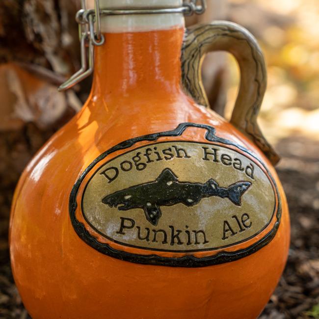 An Orange Punkin Growler by Romanick Pottery, with a handle, labeled "Dogfish Head Punkin Ale" and a fish illustration, sits outdoors on the ground.