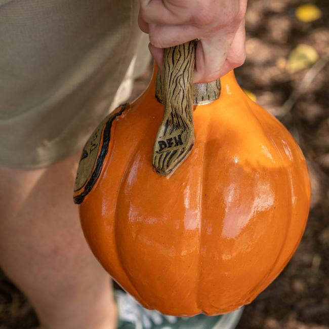 A person in khaki shorts holds the Orange Punkin Growler by Romanick Pottery, a ceramic pumpkin with a textured brown stem labeled DEN, set amid dirt and leaves—evoking the charm of unique handmade growlers.