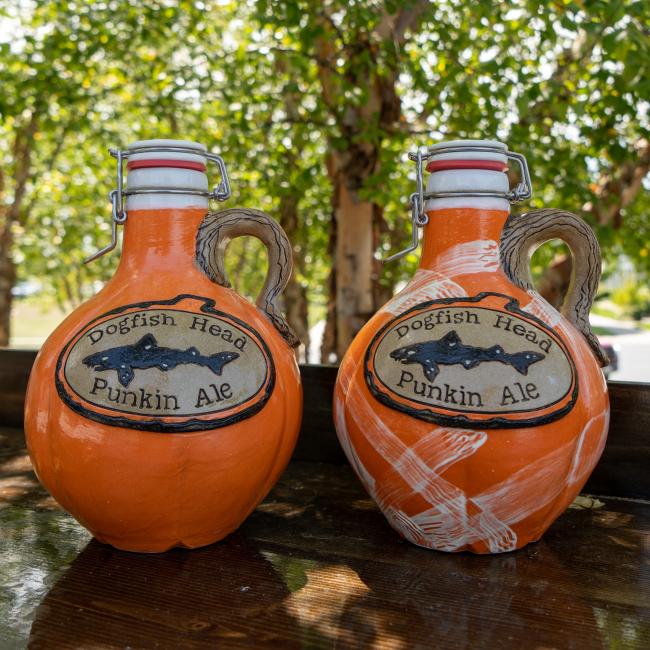 Two Orange Punkin Growlers by Romanick Pottery, each with a handle and flip-top lid, rest on a wooden outdoor surface with blurred green trees in the background.