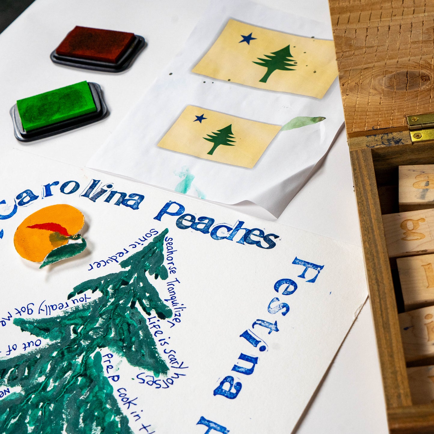 Close-up of Festina Peche screen print poster in progress with green and orange inks, rubber stamps, and carved print blocks on a studio work surface