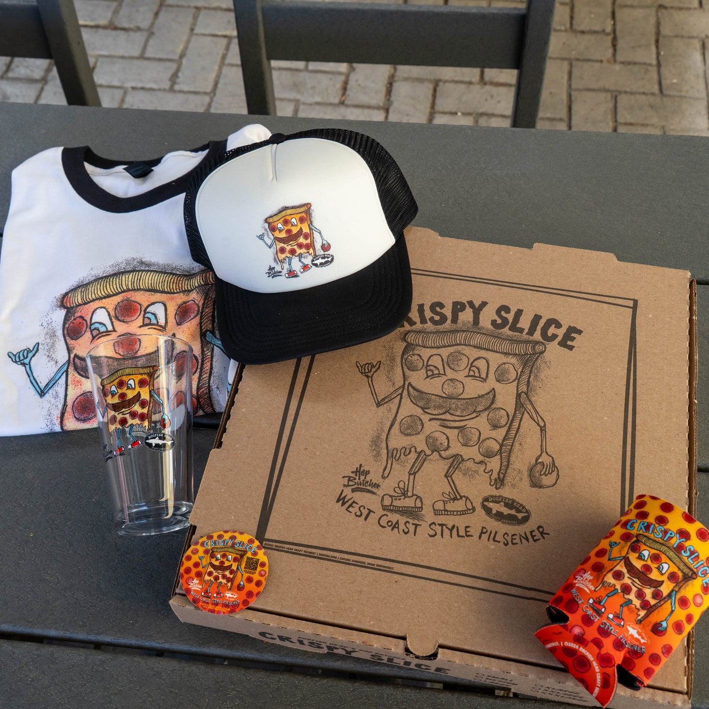 The Crispy Slice Merch Bundle by GO WEST T-SHIRT COMPANY includes a tee, trucker hat, pint glass with a pizza character, pizza box with the same design, and a colorful can cooler.