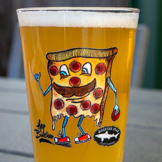 A clear pint glass filled with golden beer, featuring a sketch pizza slice character with blue arms and legs, red pepperoni slices, and a black circular logo that reads Dogfish Head Craft Brewery. The glass is photographed on a gray outdoor table with a blurred background.