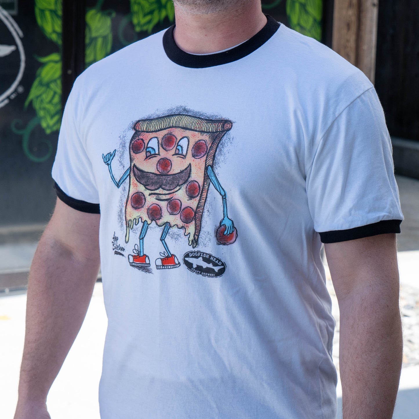 A person wears the GO WEST T-SHIRT COMPANY Crispy Slice Tee Shirt, a white ringer tee with black trim featuring a cartoon pizza slice with a smiling face, arms, legs, and sneakers, standing outdoors.
