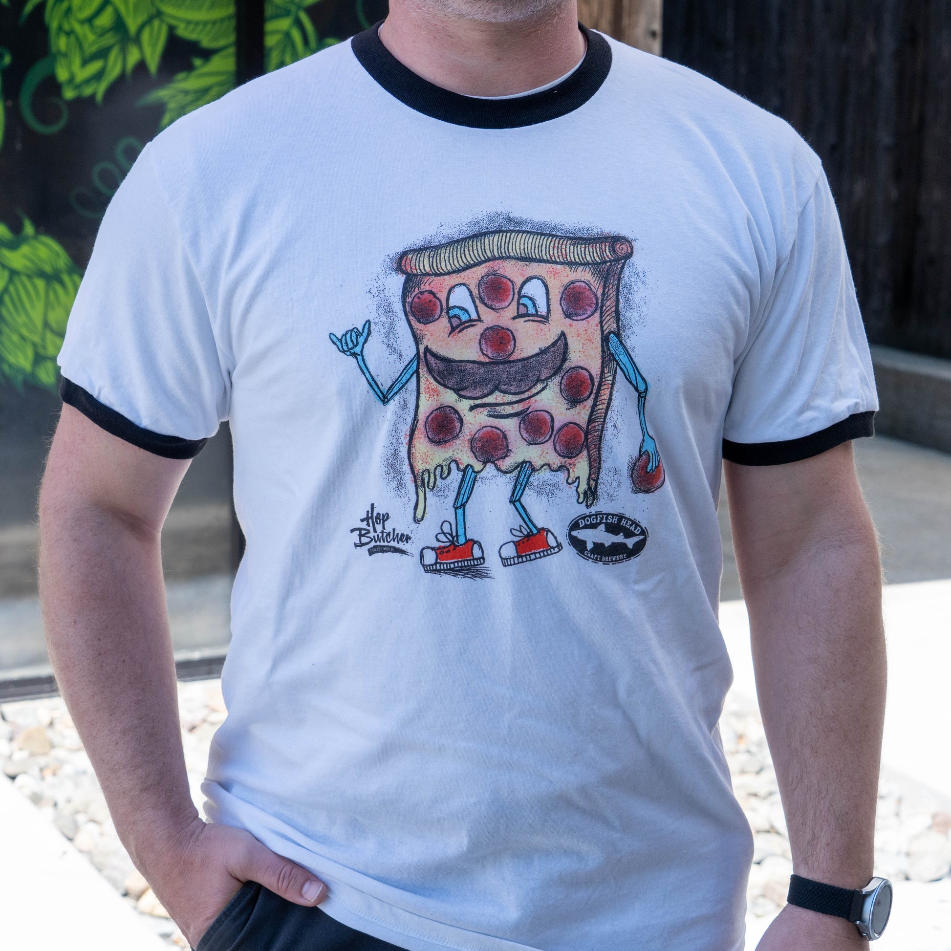 A person wears the Crispy Slice Tee Shirt by GO WEST T-SHIRT COMPANY—a white ringer tee featuring a cartoon pepperoni pizza slice with arms, legs, sneakers, skateboard, and hand gesture—set against green leaves and a wooden fence.