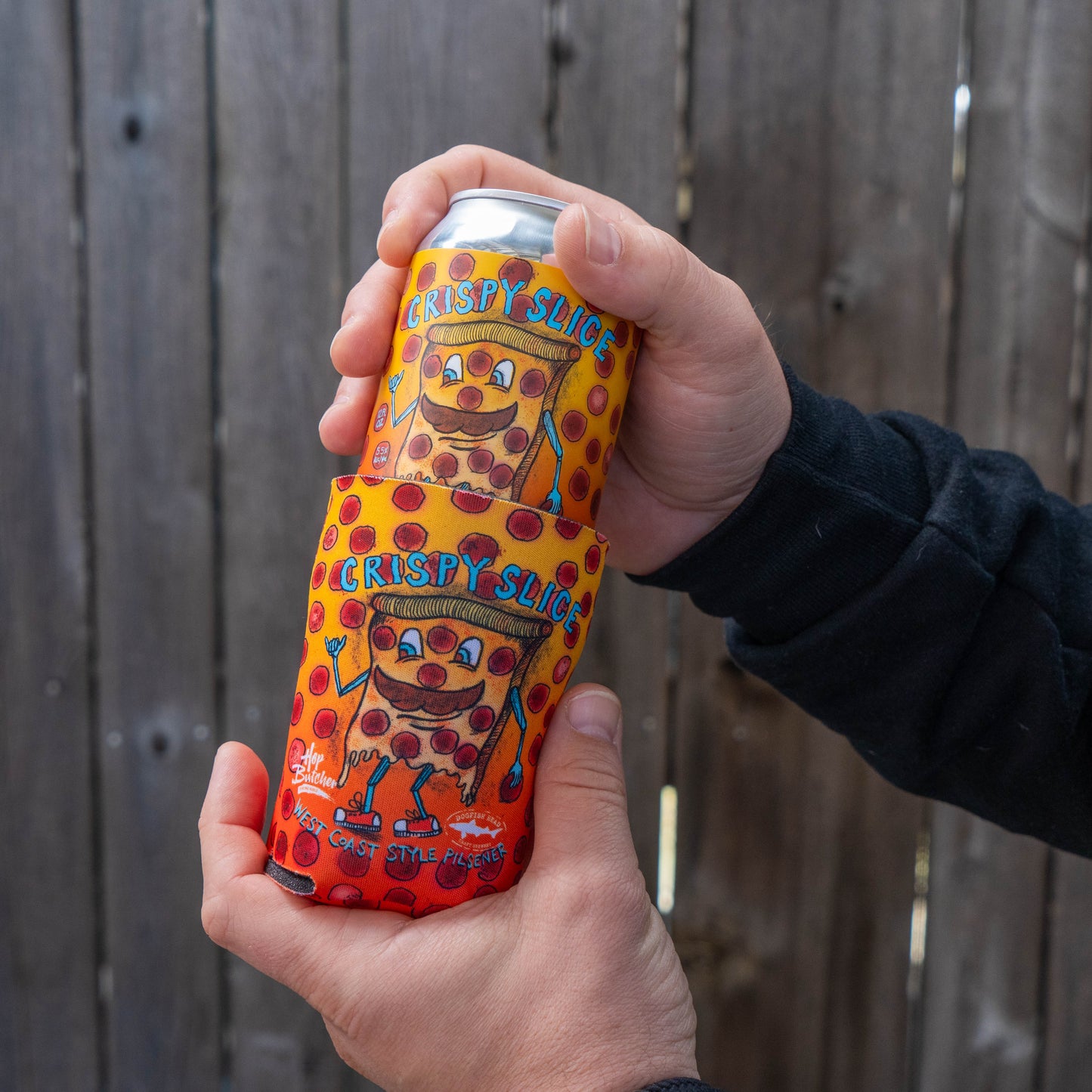 Two hands hold a beer can featuring the Crispy Slice pizza design from the GO WEST T-SHIRT COMPANY’s Crispy Slice Merch Bundle, with a matching can cooler resting on top.