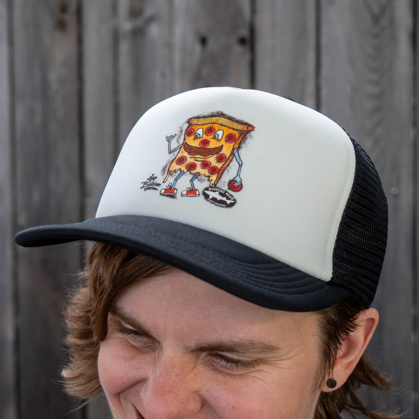 A smiling person holds a matching tee while wearing the GO WEST T-SHIRT COMPANY Crispy Slice Merch Bundle: a black and white trucker hat with a cartoon pizza slice with boxing gloves, in front of a wooden fence.