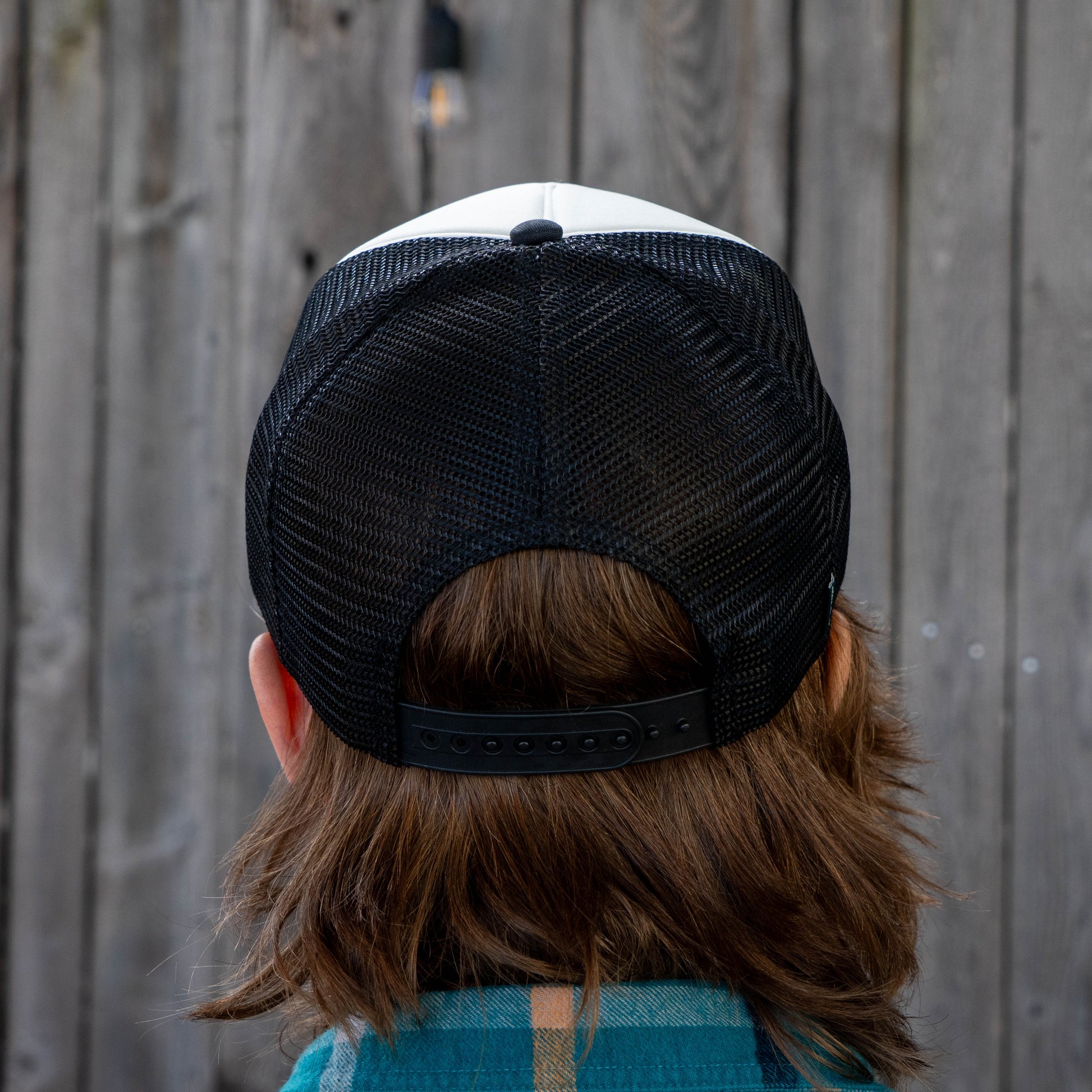 A person with medium-length brown hair wears a GO WEST T-SHIRT COMPANY Crispy Slice Merch Bundle black mesh baseball cap backwards and a casual tee, standing in front of a wooden fence.