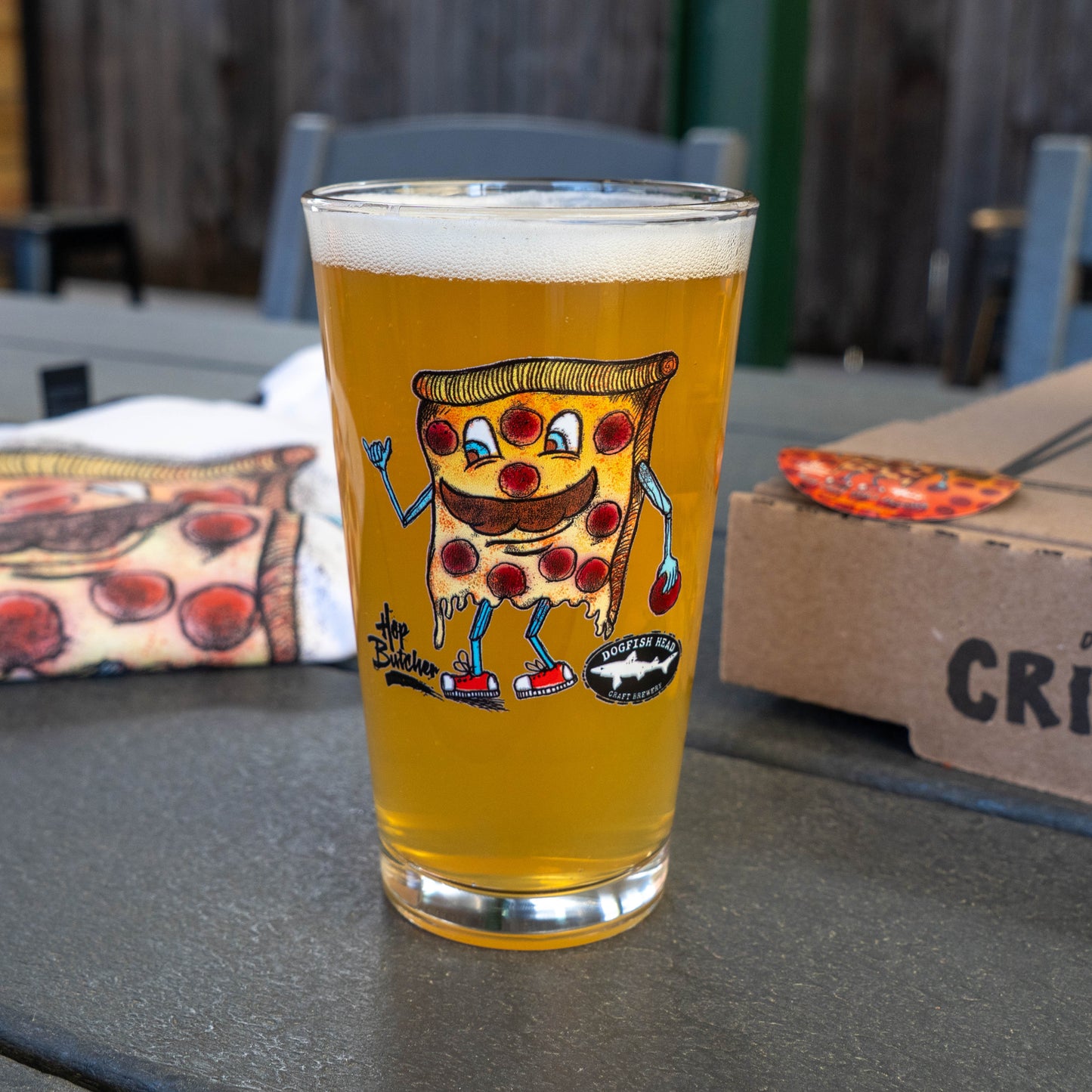 A pint glass from the Crispy Slice Merch Bundle by GO WEST T-SHIRT COMPANY sits on a table, filled with light beer. The glass shows a cartoon pizza slice; behind it are a pizza box, themed cloth, and matching can cooler.
