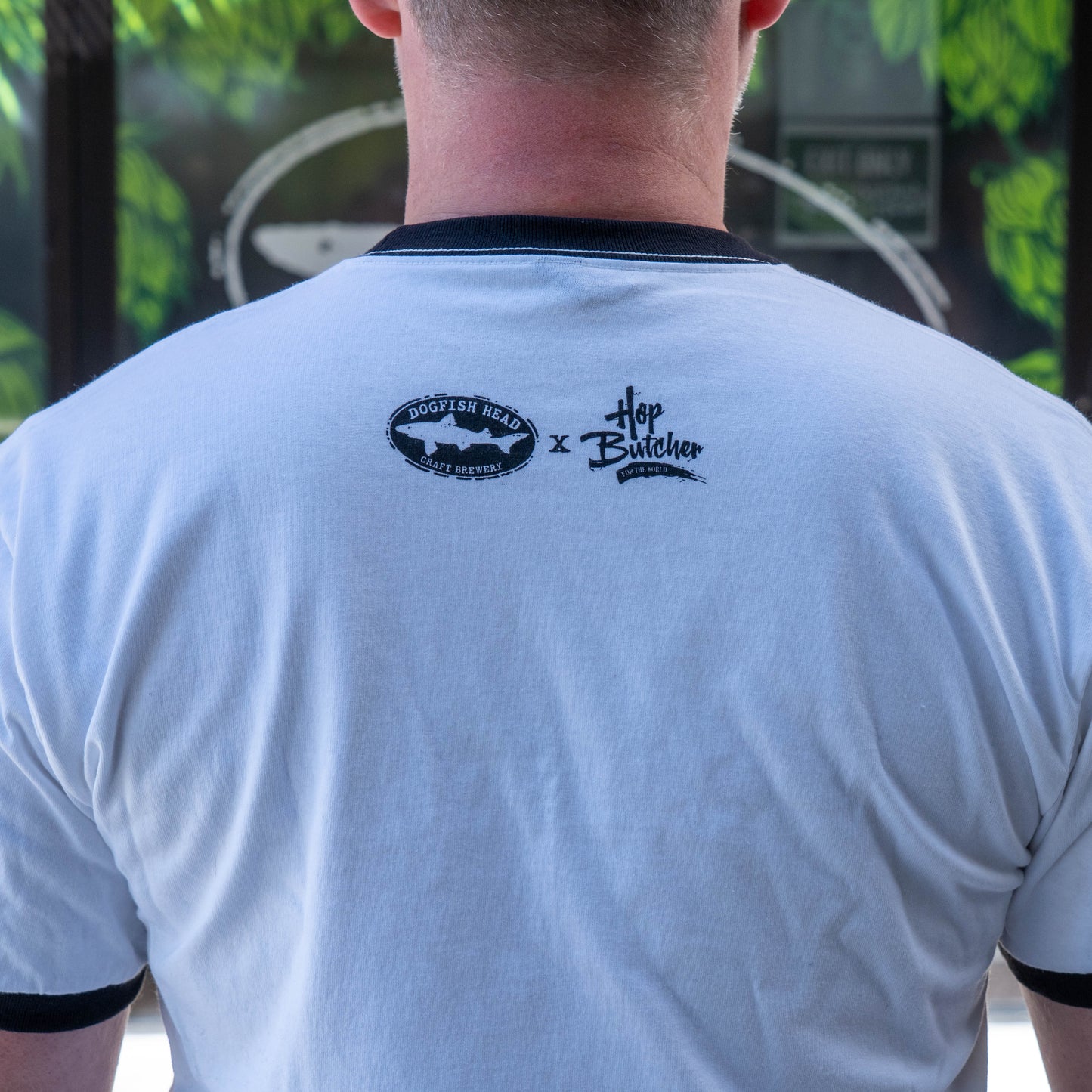 A person wears a GO WEST T-SHIRT COMPANY Crispy Slice Merch Bundle white tee with black trim, showing the Dogfish Head and Hop Butcher logos on the back. Green leaves and a white circle blur in the background near a pint glass.