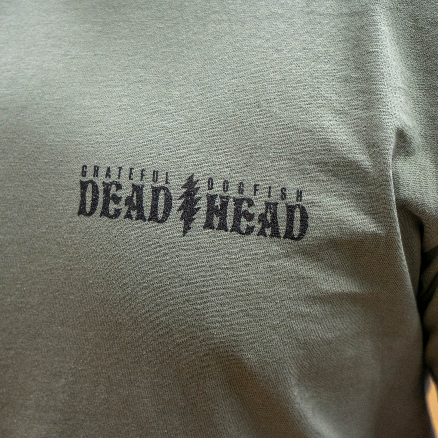 Close up of the front view of Dogfish Head x Grateful Dead Citrus Daydream Tee in the color sand. The front left chest has a logo in a dark grey color reading "Grateful Dead, Dogfish Head" with a grateful dead lightning bolt symbol dividing the two logos. 