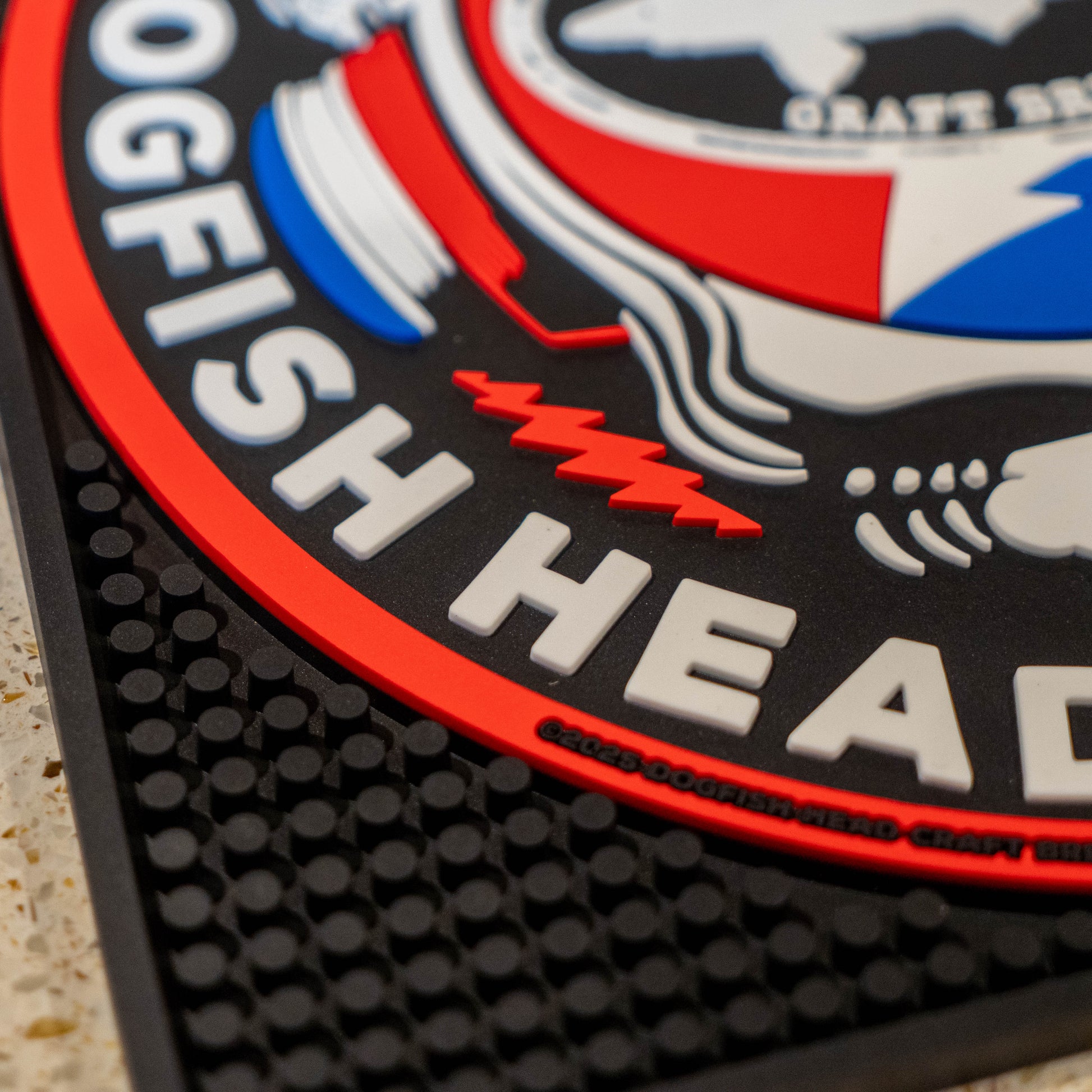 Close up of black square bar mat with a red, white, and blue Dogfish Head x Grateful Dead Logo.