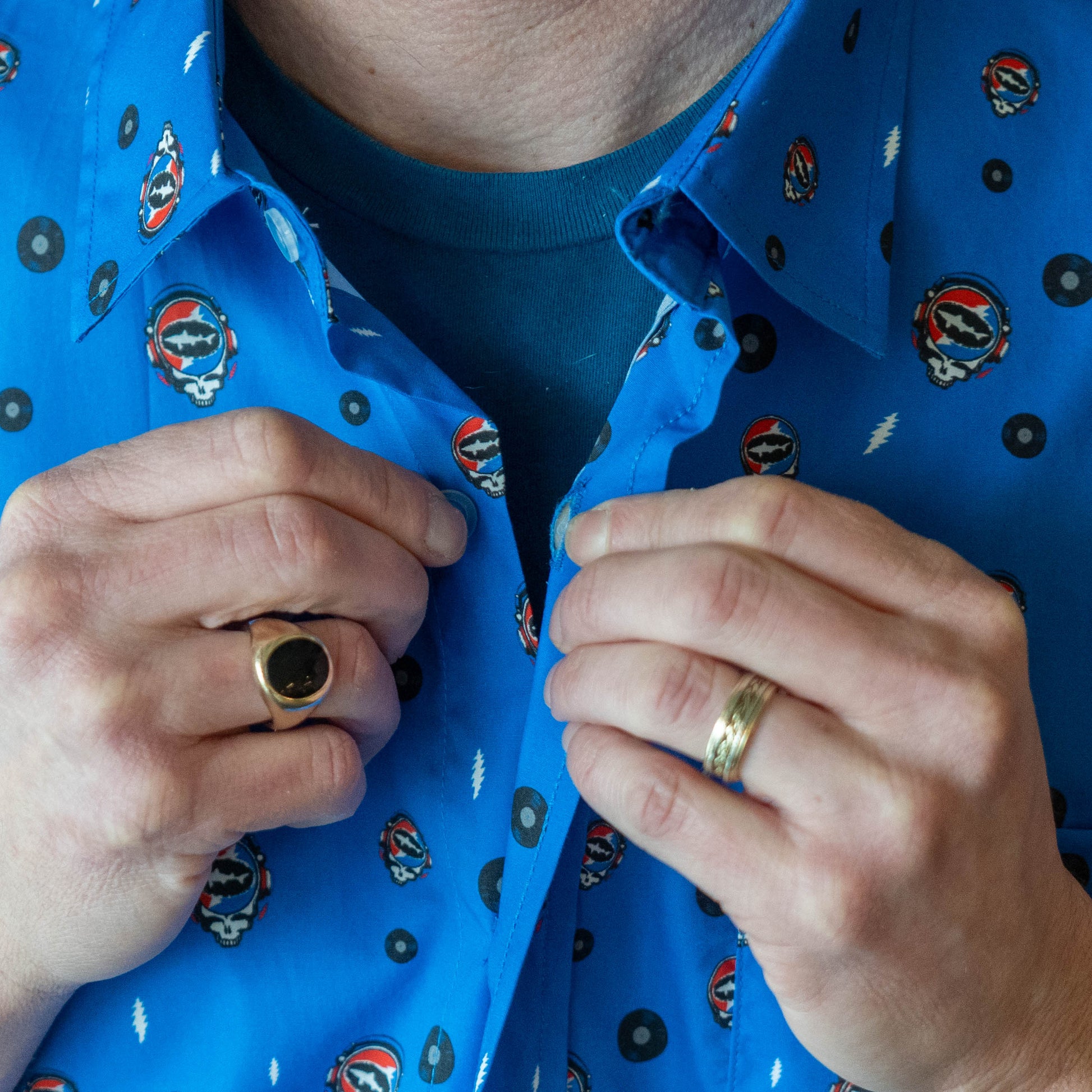 Close up of a person buttoning up a blue Dogfish Head x Grateful Dead button-down shirt, designed in small lightning bolts, records, and Dogfish Head x Grateful dead red, white, and blue logos.