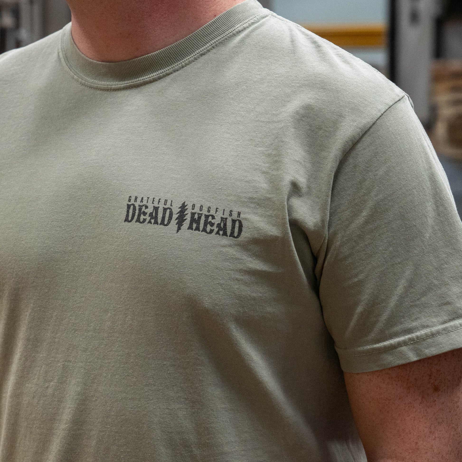 Close up of the front view of Dogfish Head x Grateful Dead Citrus Daydream Tee in the color sand. The front left chest has a logo in a dark grey color reading "Grateful Dead, Dogfish Head" with a grateful dead lightning bolt symbol dividing the two logos. 