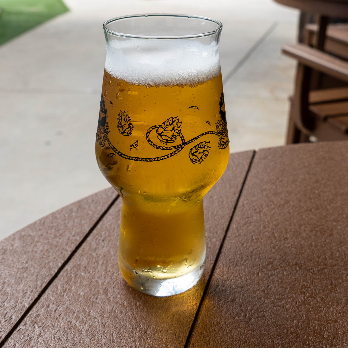 An Anchor IPA Glass filled with golden beer and foam, featuring a black graphic of hop cones and vines, sitting on a wooden table.