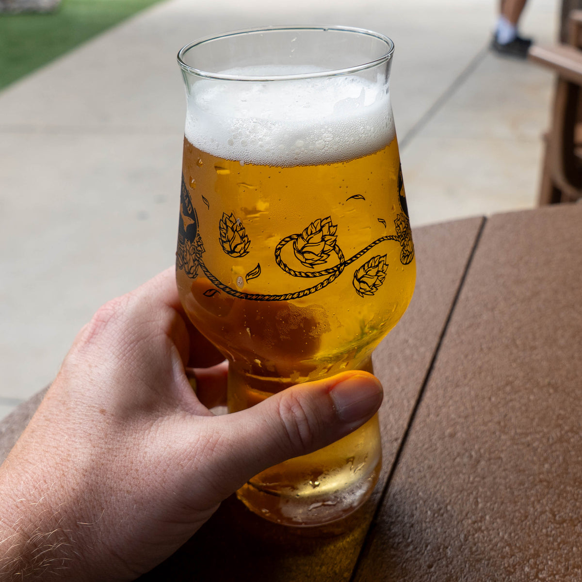 A hand holds an Anchor IPA Glass filled with golden ale topped with foam, featuring a black illustration of hop vines and cones.