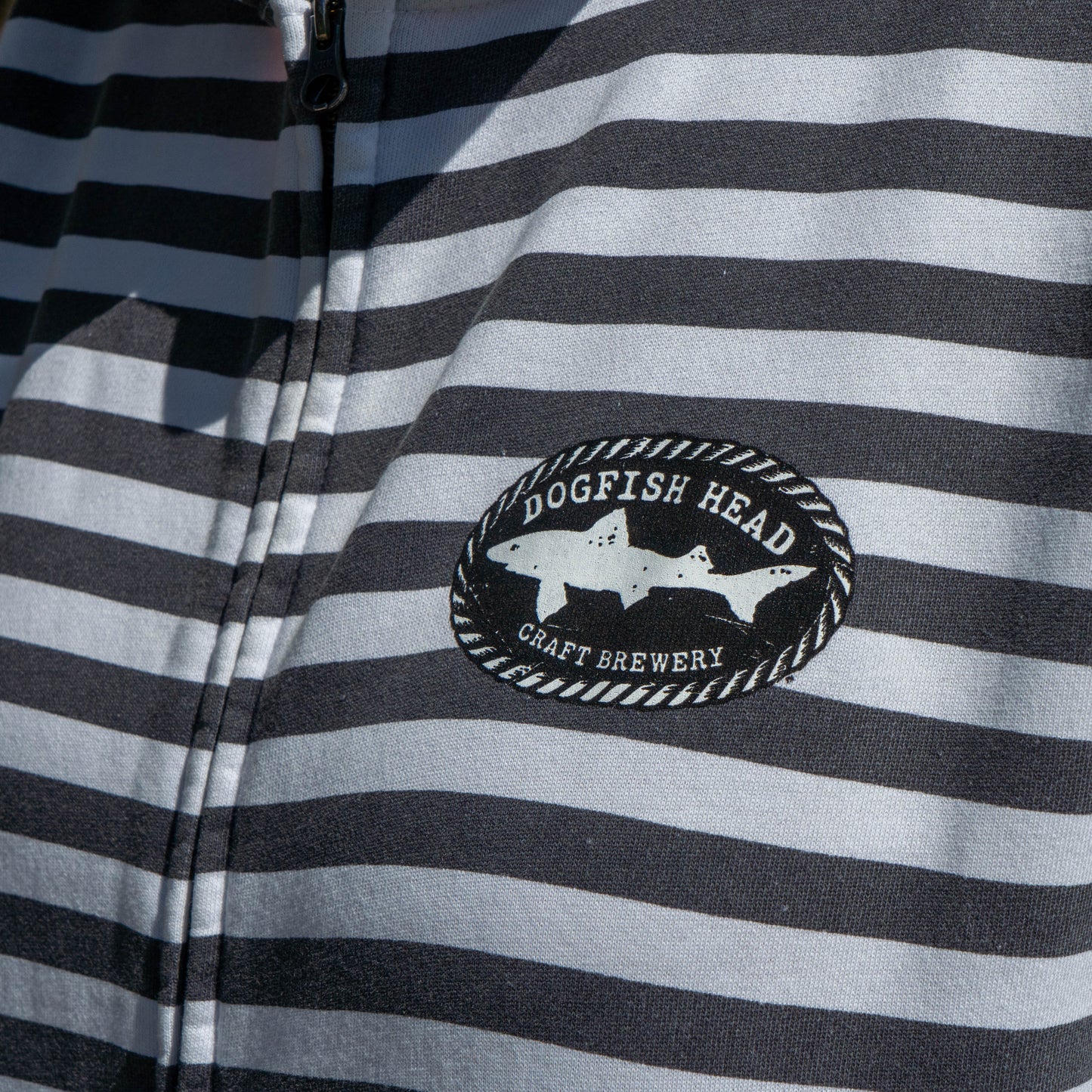 Close-up of a black and white horizontally striped zip-up jacket featuring a circular 'Dogfish Head Craft Brewery' shark patch on the chest.