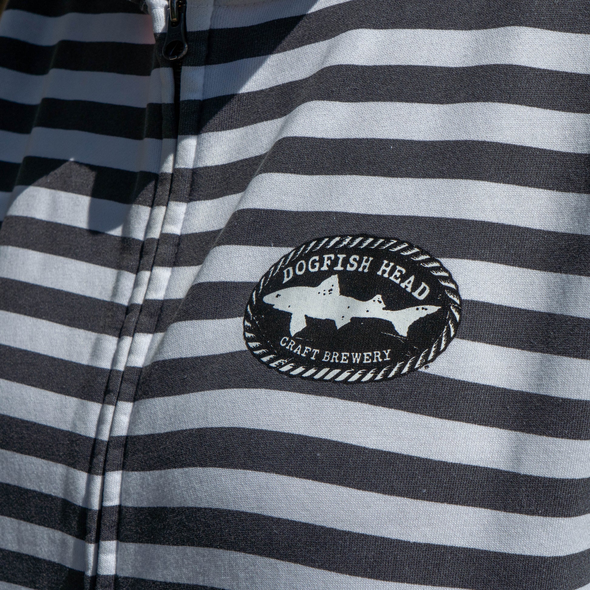 Close-up of a black and white horizontally striped zip-up jacket featuring a circular 'Dogfish Head Craft Brewery' shark patch on the chest.
