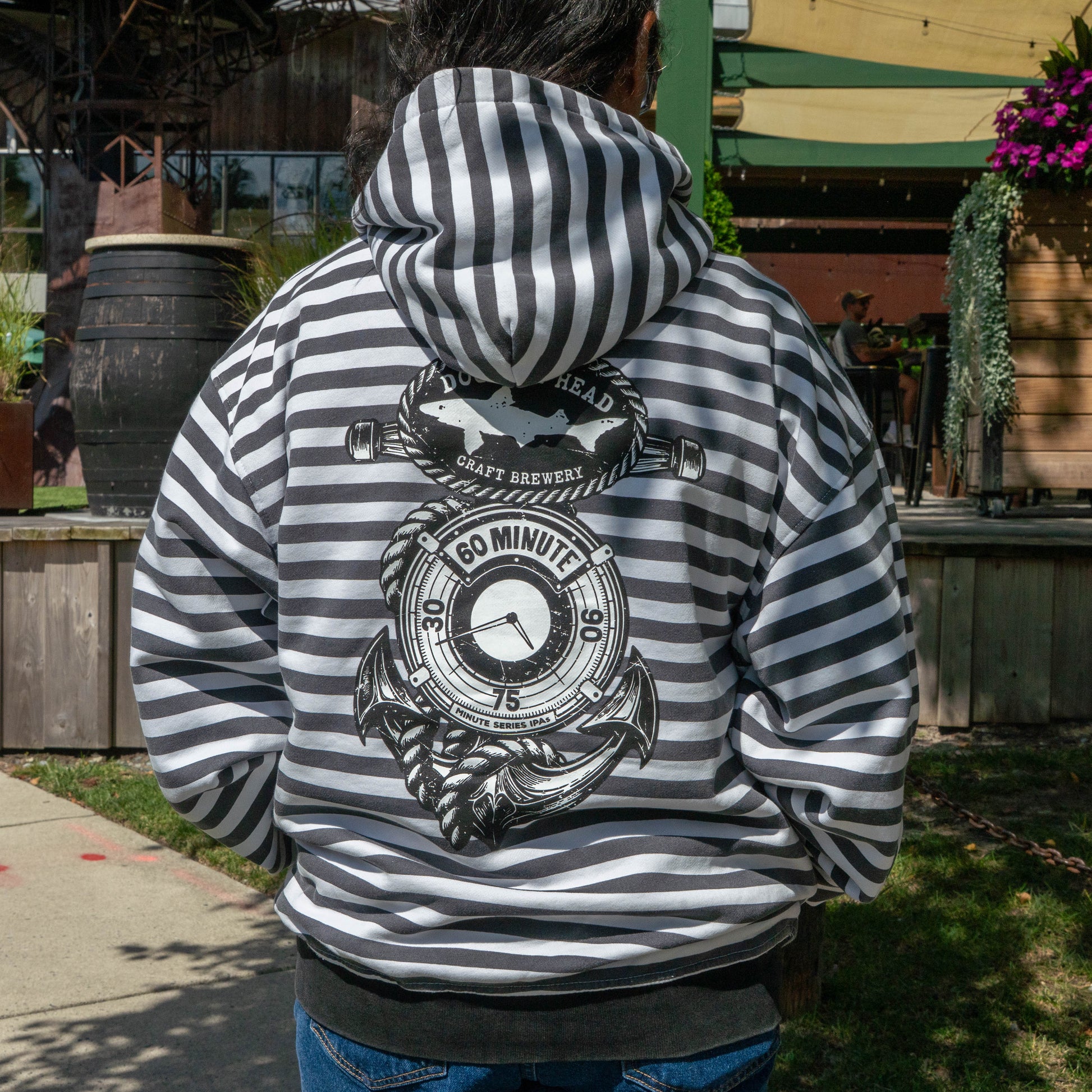 A person wearing the GO WEST T-SHIRT COMPANY B&W Striped Zip Hoodie, which features a nautical-themed 60 Minute craft brewery design on the back, is standing outdoors near wooden railings and plants.