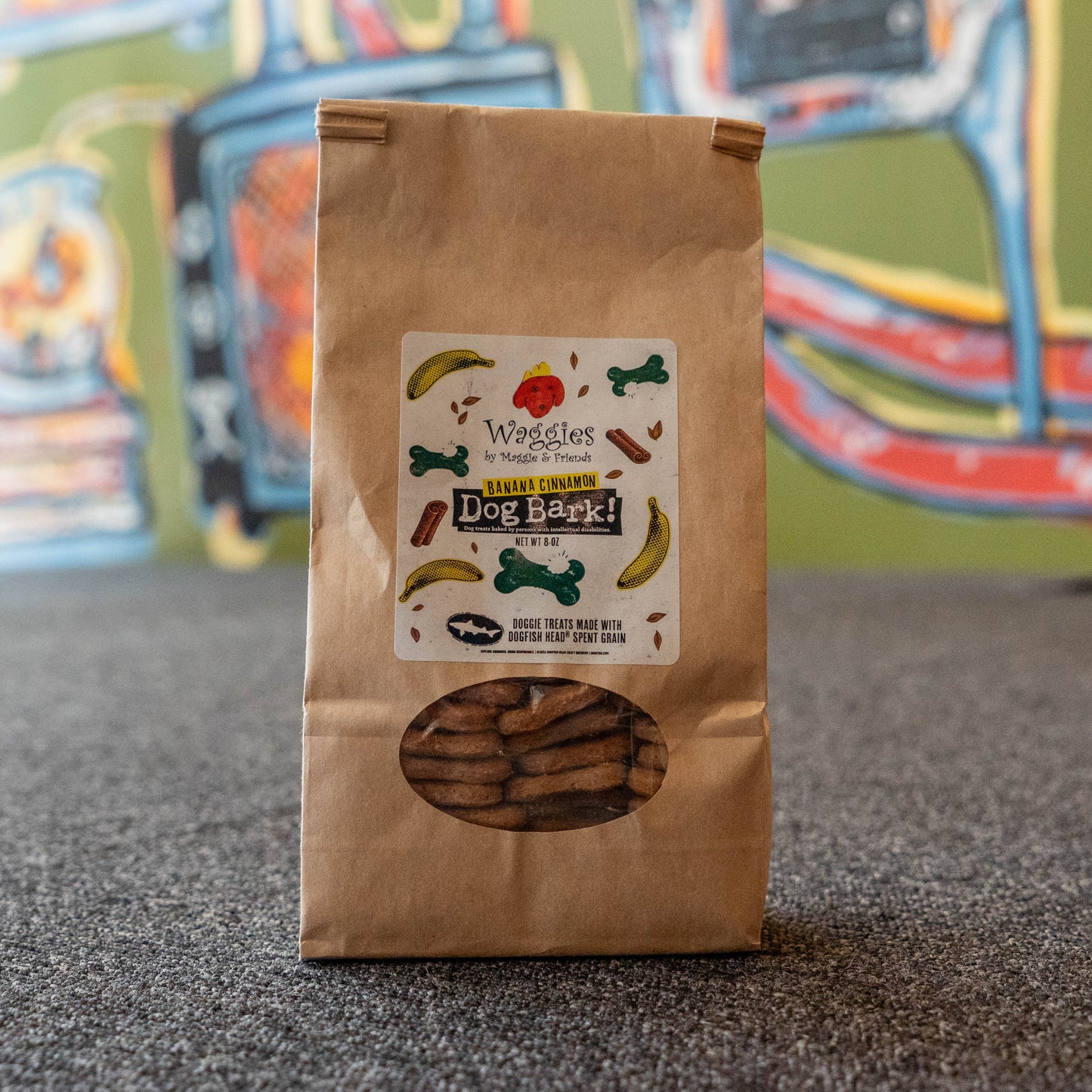 A brown paper bag of Waggles by Waggies Natural Treats Dog Bark! treats, featuring a clear window showing the brown bark treats inside, set on a gray carpet with a colorful blurred mural in the background.