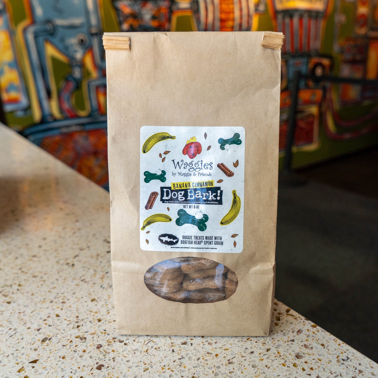 A brown kraft paper bag of Waggies Pet Treats 'Dog Bark' treats. featuring a clear window at the bottom showing the treats inside, set on a speckled countertop with a colorful blurred background.