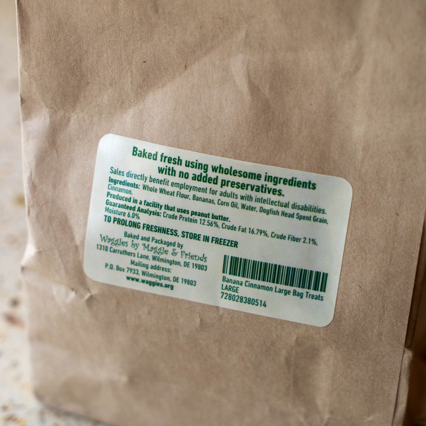 A close-up of a brown kraft paper bag with a white product label. The label reads 'Baked fresh using wholesome ingredients with no added preservatives.