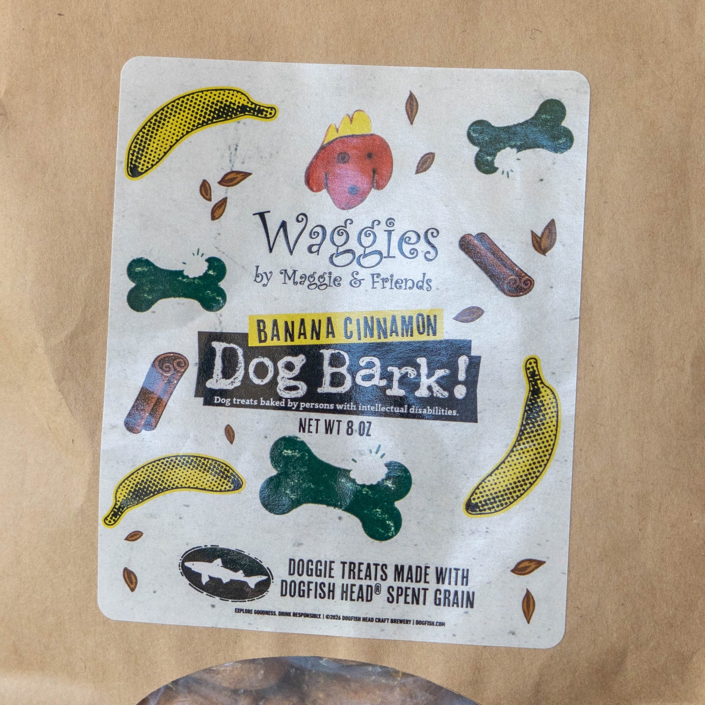 A package of Waggies by Maggie & Friends Dog Bark! Banana Cinnamon dog treats, featuring a white label with colorful illustrations of dog treats and leaves, placed on a brown paper bag.
