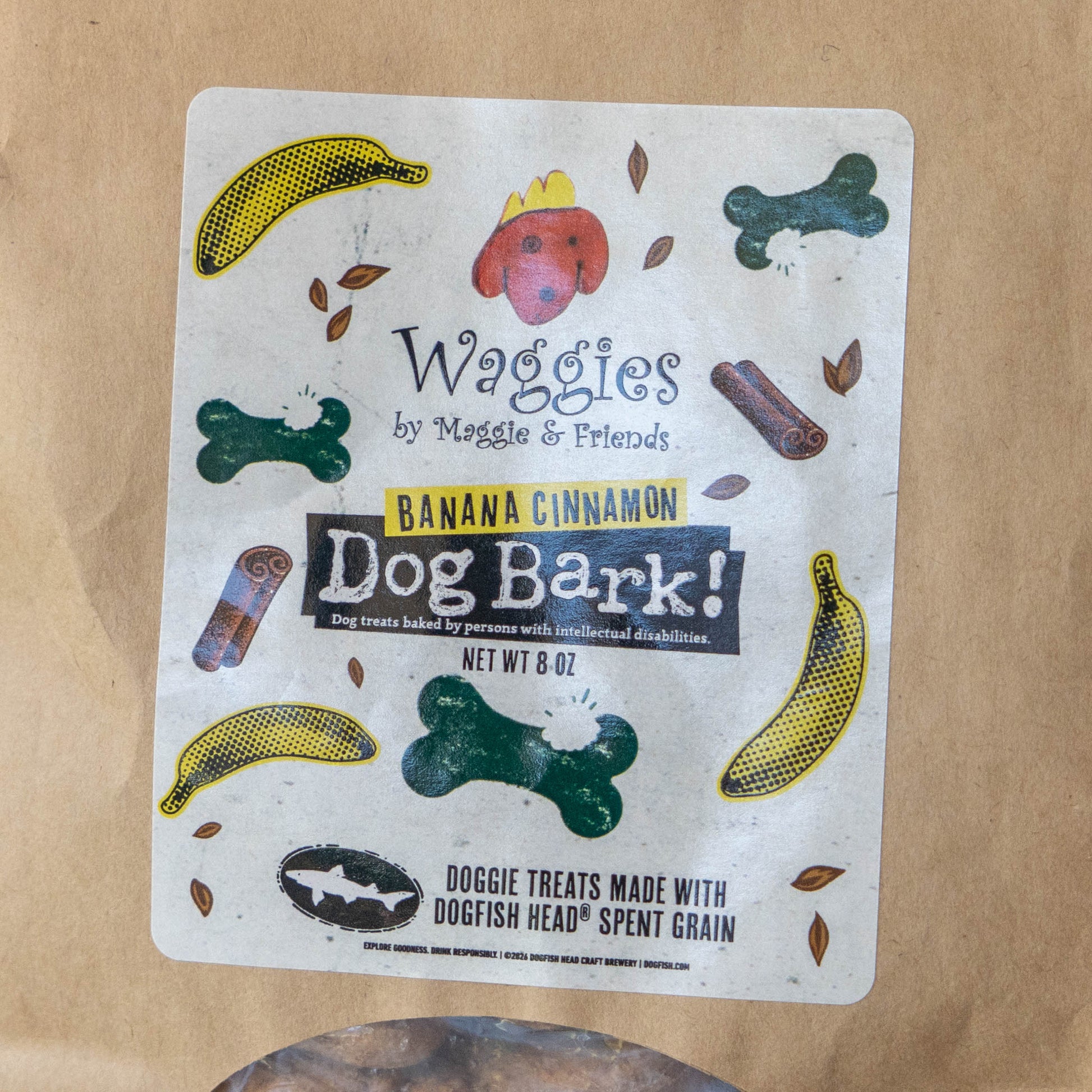 A package of Waggies by Maggie & Friends Dog Bark! Banana Cinnamon dog treats, featuring a white label with colorful illustrations of dog treats and leaves, placed on a brown paper bag.