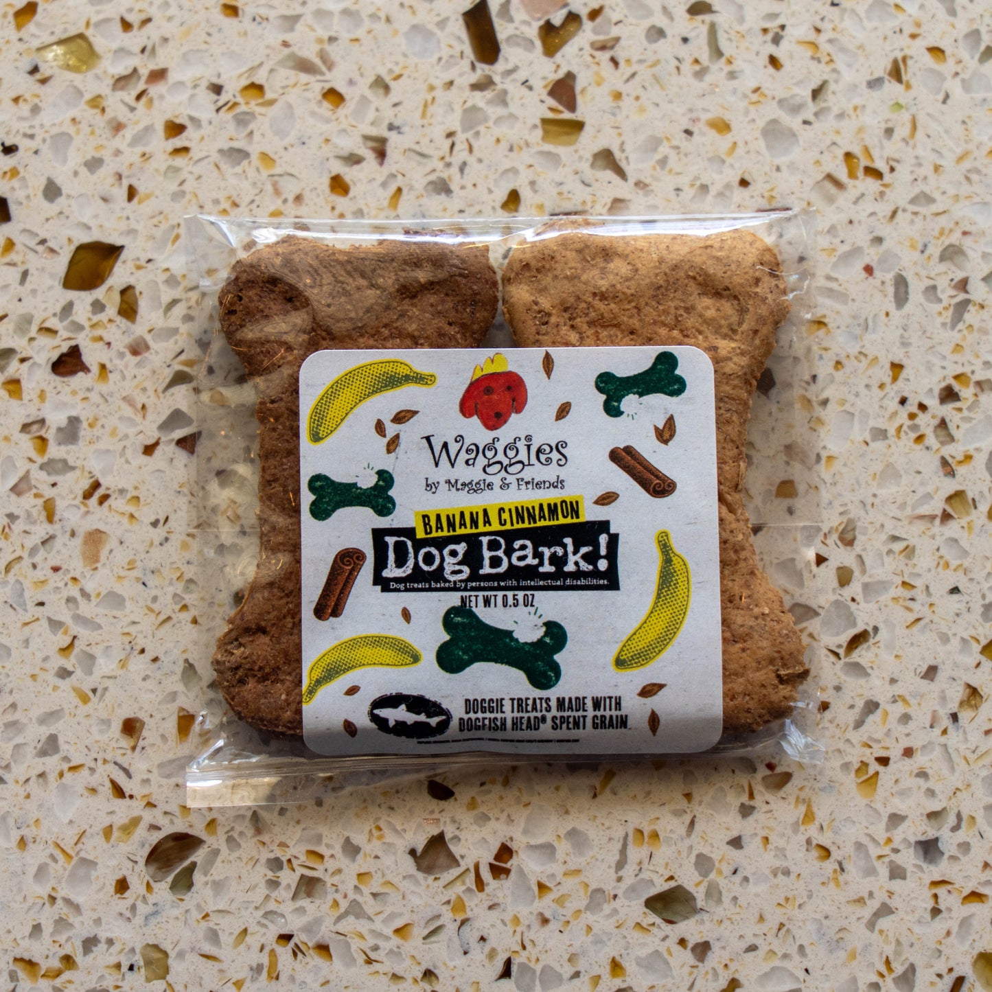 A package of Waggies by Waggies 'Banana Cinnamon Dog Bark!' dog treats, shaped like bone-shaped treats, placed on a speckled beige countertop.