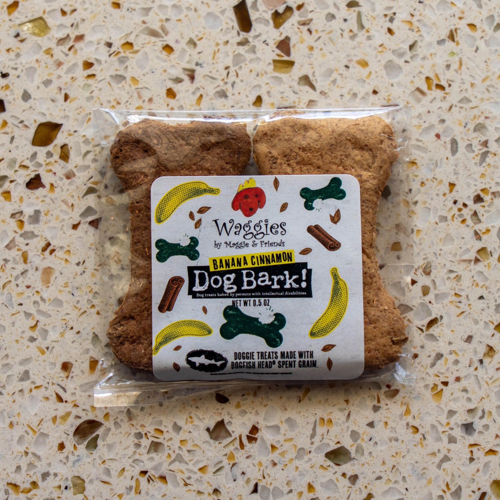 A package of Waggies by Waggies 'Banana Cinnamon Dog Bark!' dog treats, shaped like bone-shaped treats, placed on a speckled beige countertop.