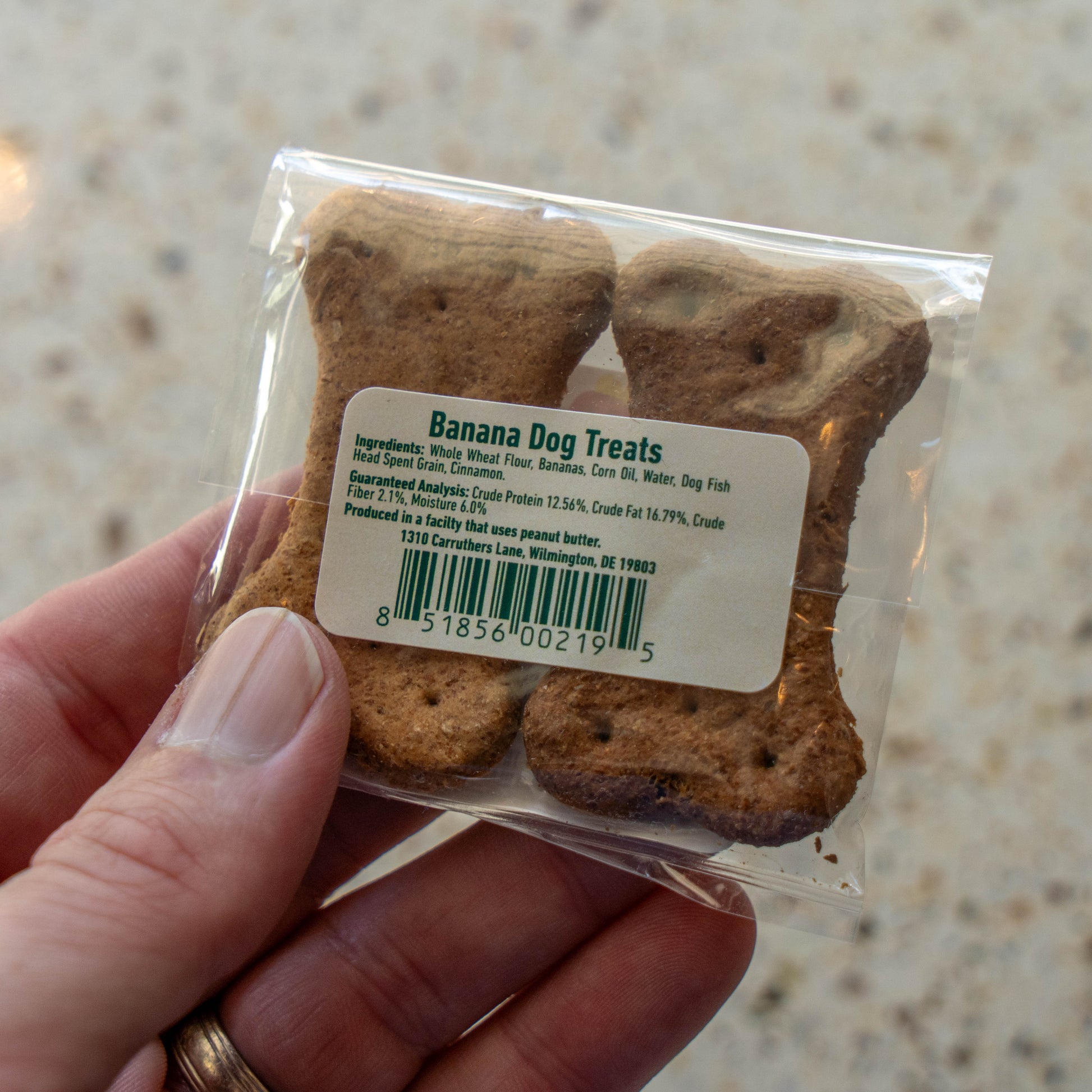 A hand holding a clear plastic bag of bone-shaped banana dog treats with a white label, set against a light beige speckled countertop background.